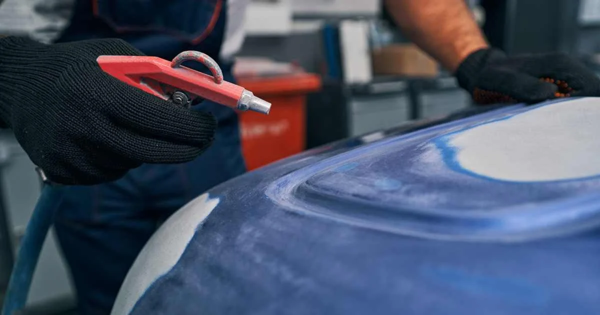 Worker using a red air gun to clean or blow off a blue car hood surface.